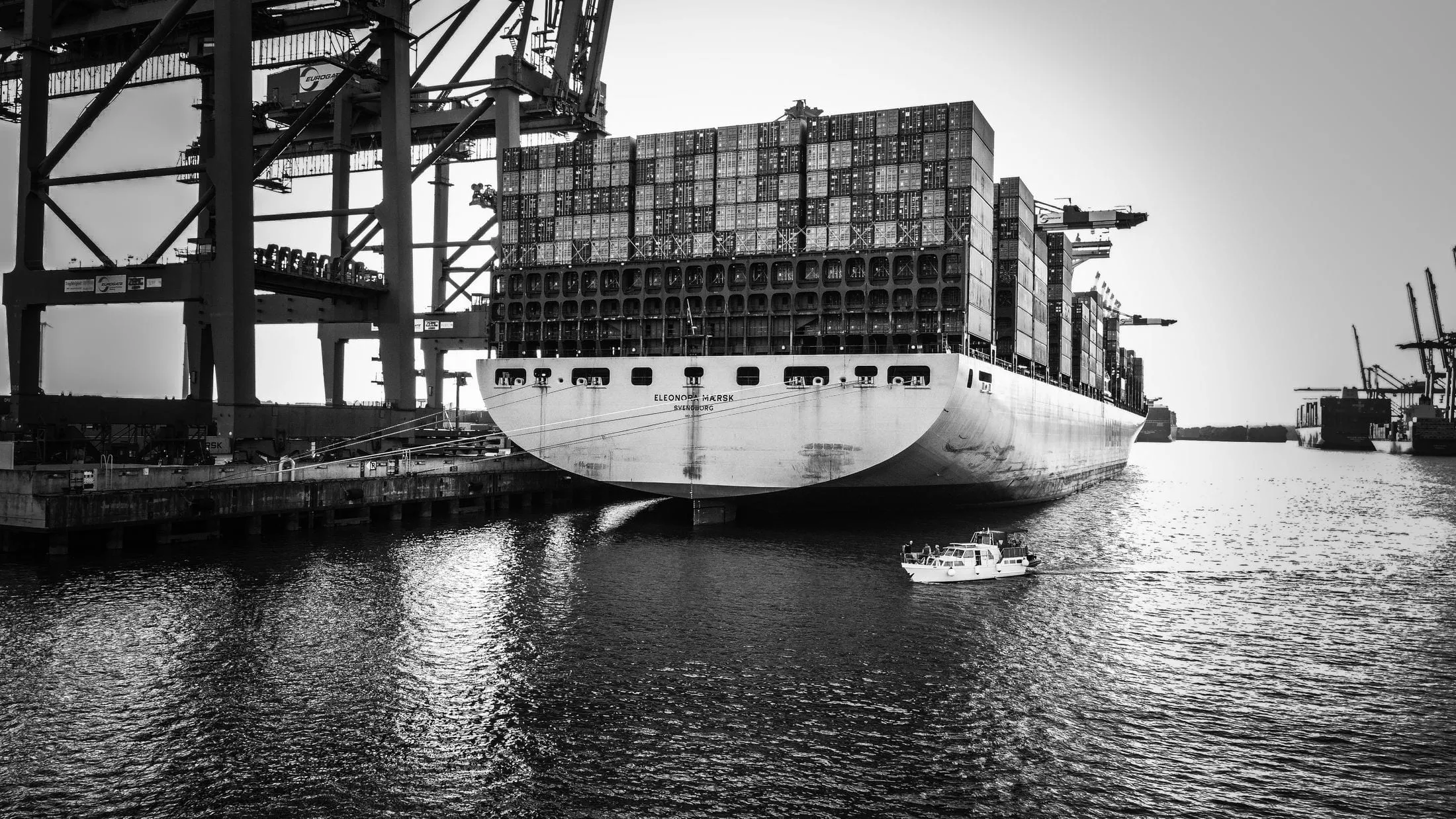 Black and white photo of a loaded container ship berthed under a gantry crane