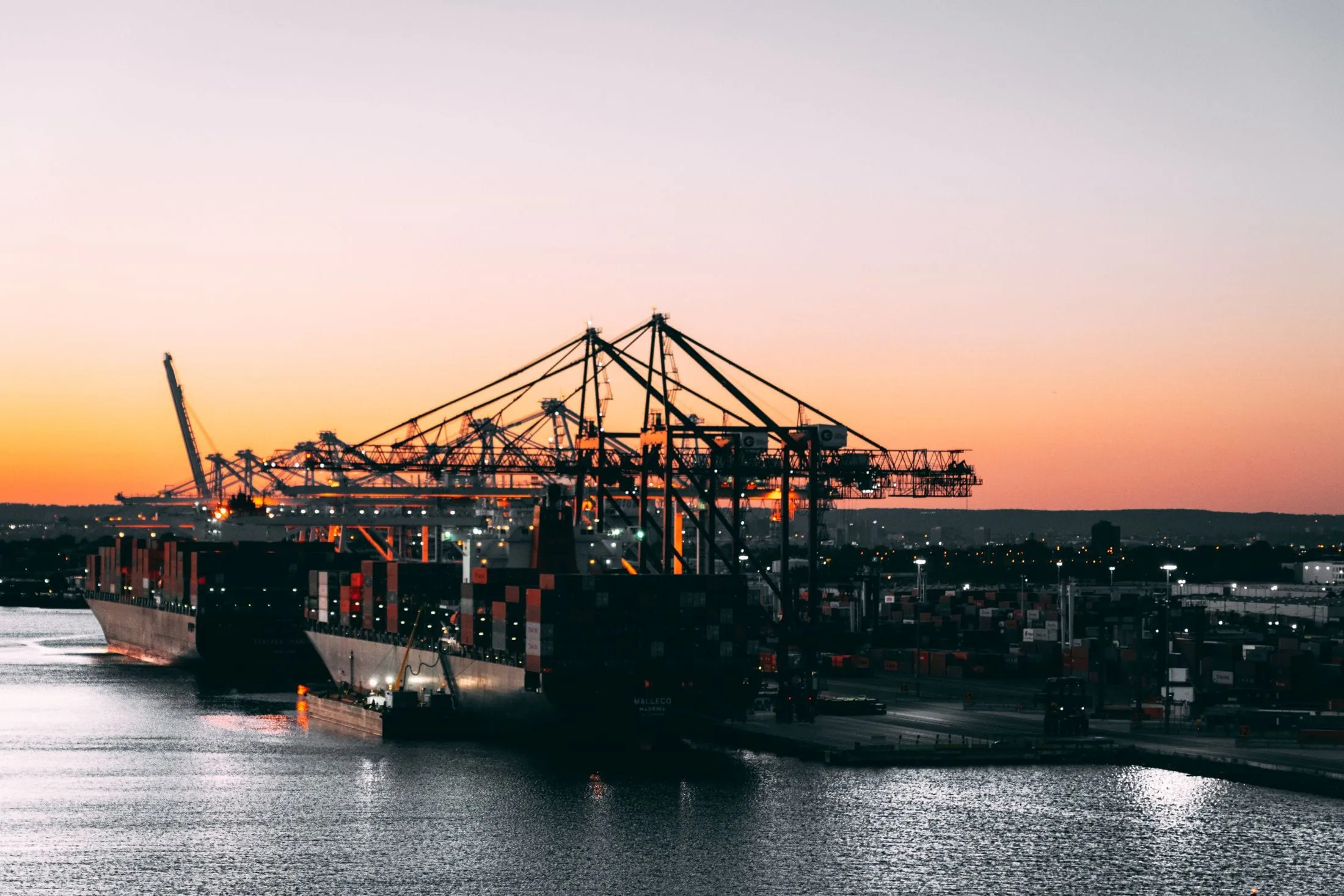 Container ships under gantry cranes at a port at dusk, orange sky behind